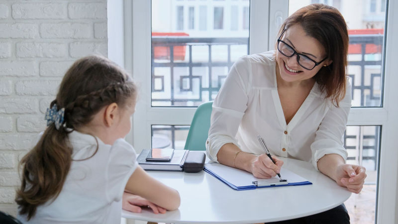 smiling woman takes notes on a clip board while sitting at a small table with a child