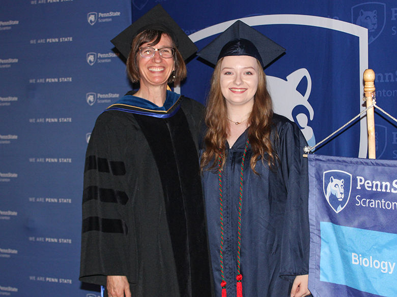 a smiling graduate in a cap and gown stands next to smiling biology professor in cap and gown