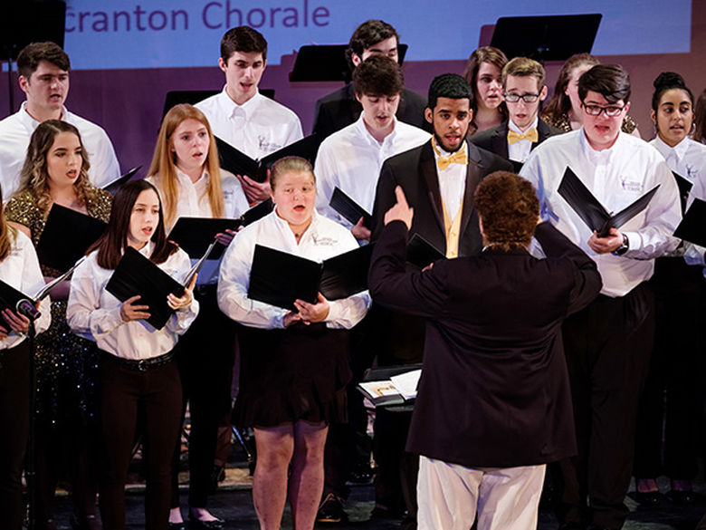 a conductor in front of a 15-person choir reading sheet music and singing