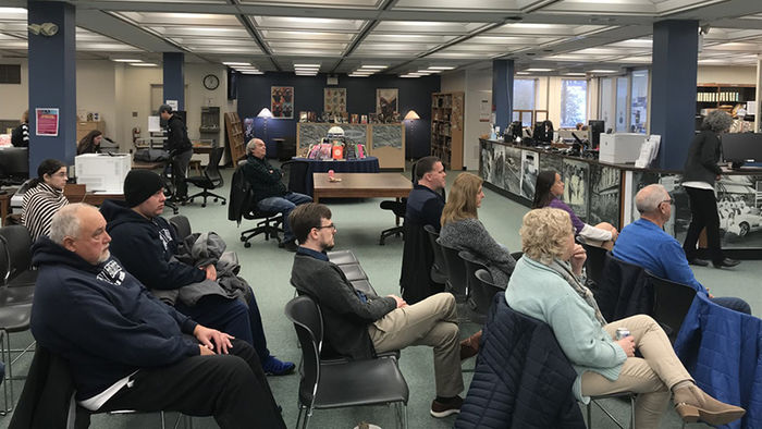 large group of people sitting in rows of chairs in a library listening to a speaker