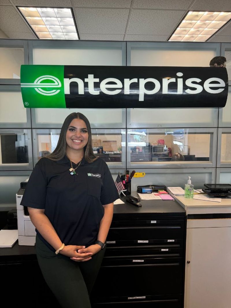 A student stands in front of a desk with an Enterprise logo in the background