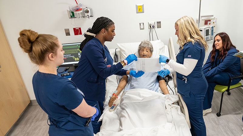 four students in scrubs stand around an aged mannequin in a lab that looks like a hospital room