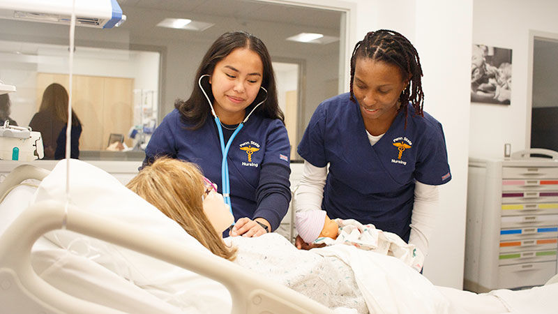 two students in scrubs take the vital signs of a mannequin in a lab that looks like a hospital room