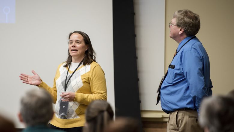 Gina speaks while Griff listens and looks toward presentation screen