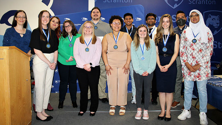 12 students with medals around their necks pose with smiling teacher