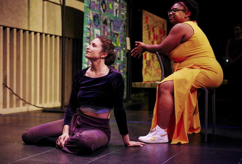 A white woman sits straight on a floor stage next to a Black woman sitting in a chair.